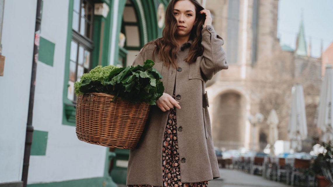 A woman in a brown coat carries a wicker basket of green vegetables while strolling through Erfurt, Germany.