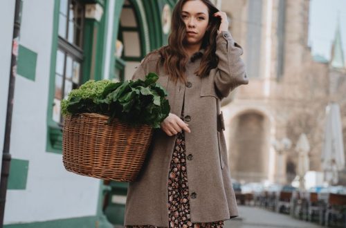 A woman in a brown coat carries a wicker basket of green vegetables while strolling through Erfurt, Germany.