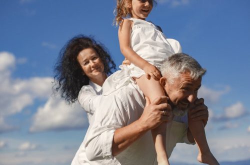 Grandparents playing with granddaughter outdoors under bright blue sky.