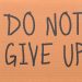 Cardboard sign reading 'Do Not Give Up' placed on green turf, symbolizing perseverance.